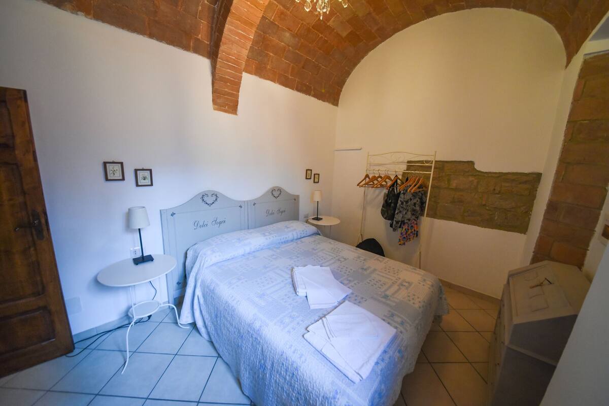 A bedroom with a bed, a bedside table, a lamp, a hanging clothes rack, and a brick wall.