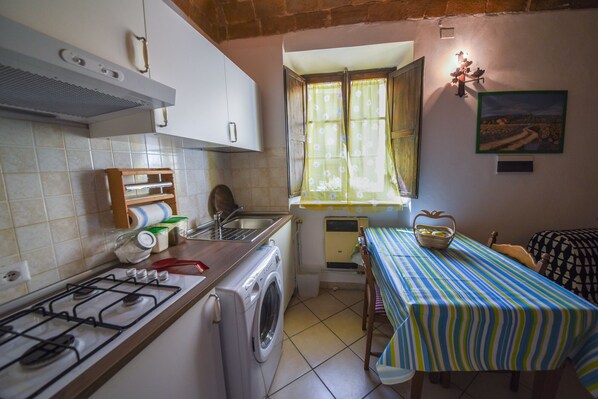 A compact kitchen with a gas stove, a washing machine, and a small dining area with a striped tablecloth. - Il Sogno di Annalisa (Volterra)