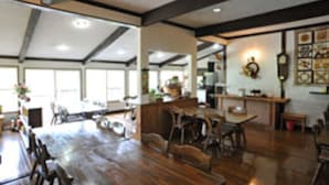 A spacious dining area with wooden tables and chairs, a kitchen in the background, and a large window providing natural light. - Pension Marionette (Hokuto)
