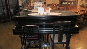 A piano with a dining table and chairs in a room with wooden flooring and a bookshelf in the background. - Pension Marionette (Hokuto)