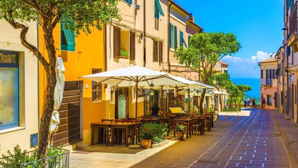A narrow street lined with outdoor cafes, featuring tables and umbrellas, and buildings with shutters and balconies. - Apartment in Capoliveri near Sandy Beaches (Capoliveri)