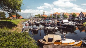 A marina with various boats docked, a wooden dock, and a backdrop of traditional buildings and a tower.