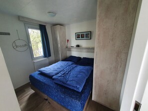 A bedroom with a bed, a window with curtains, a radiator, a framed picture on the wall, and a shelf with a small item.