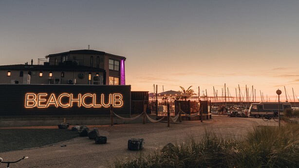A beach club with a neon sign, a building with multiple windows, and a dock area with boats.
