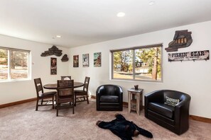 A room with a dining table, two chairs, a sofa, a small side table, and a large window with a view of trees and a house. - Goldmine Lakeview by Destination Big Bear : Luxury Lake & Mountain Views with Pool Table & Hot Tub! Walk to Village! (Big Bear Lake)
