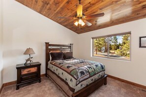 A bedroom with a wooden ceiling, a bed with a patterned quilt, a nightstand with a lamp, and a window with a view of trees. - Goldmine Lakeview by Destination Big Bear : Luxury Lake & Mountain Views with Pool Table & Hot Tub! Walk to Village! (Big Bear Lake)