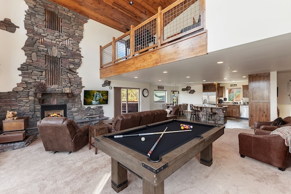 Un vaste espace de vie comprenant une table de billard, des canapés en cuir, une cheminée en pierre et un plafond haut avec des poutres en bois.
