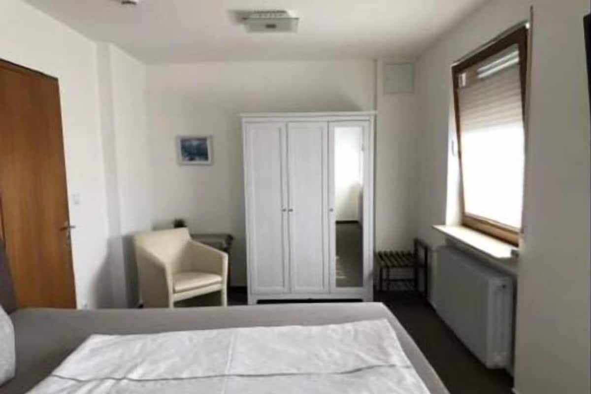 A small, well-lit bedroom with a single bed, a chair, a wardrobe, and a window with blinds.