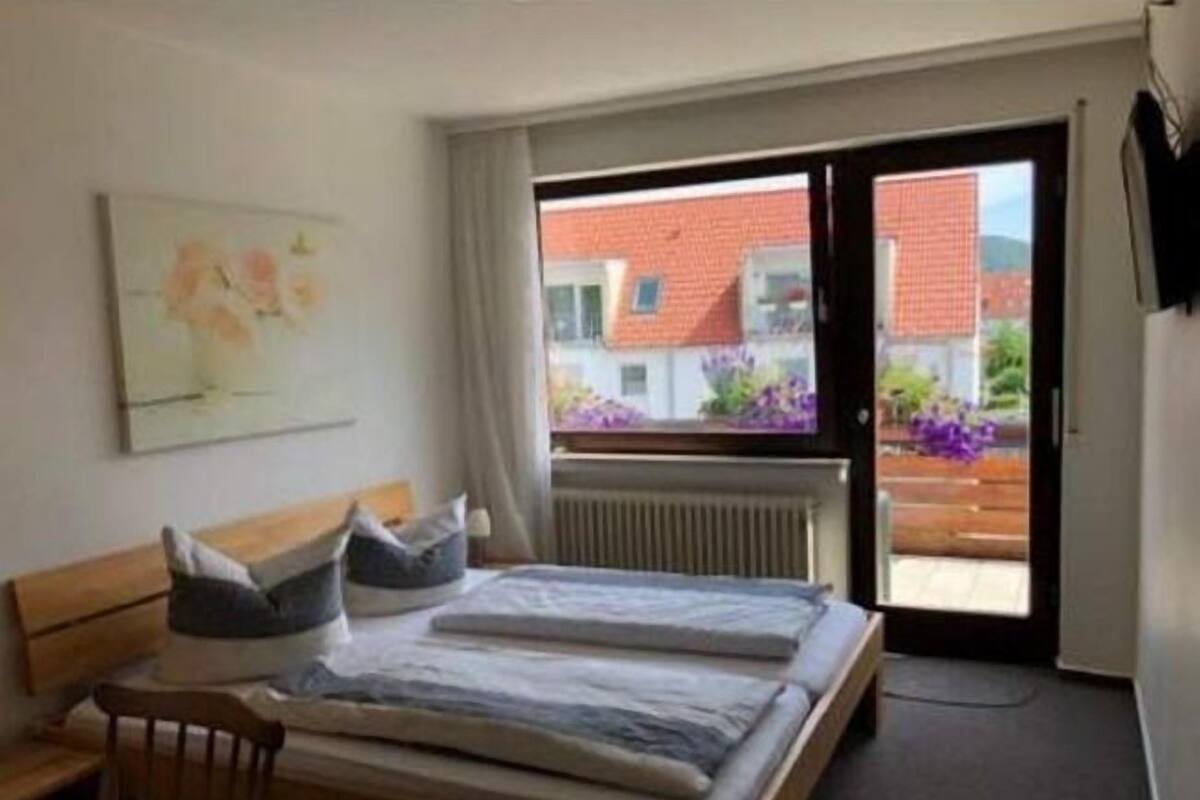 A bedroom with a bed, a chair, a radiator, and a window with a view of buildings and flowers outside.