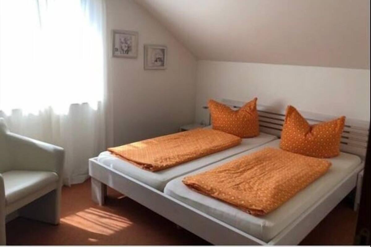 A small, single-room apartment with a bed, a chair, and a window.