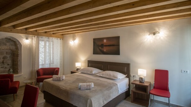 A bedroom with a large bed, two bedside tables, a nightstand, a red chair, a stone wall, and a painting on the wall. - JR Luxury Guesthouse 2 (Split)