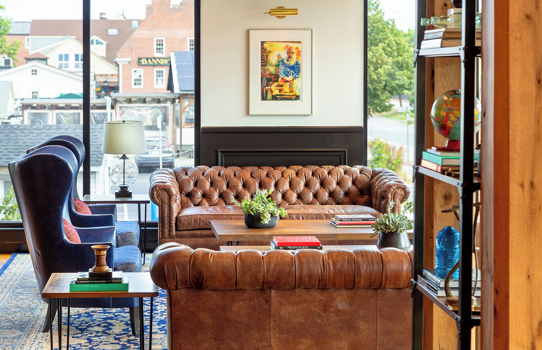 A cozy living room with a leather sofa, a blue armchair, and a coffee table with books and a plant. A framed painting hangs on the wall, and a bookshelf is visible to the right.