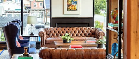 A cozy living room with a leather sofa, a blue armchair, and a coffee table with books and a plant. A framed painting hangs on the wall, and a bookshelf is visible to the right.