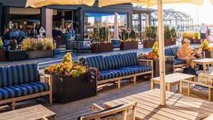 An outdoor seating area with wooden tables and chairs, blue cushioned benches, and a large umbrella providing shade.