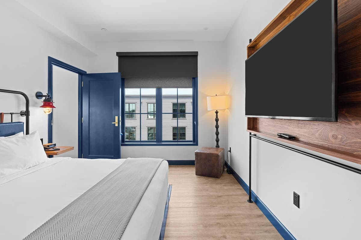 A bedroom with a bed, a desk, a chair, a TV, a lamp, and a window with a view of a building.