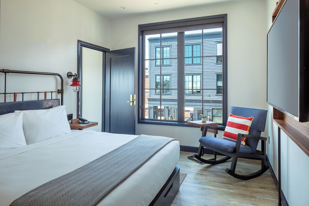 A hotel room with a bed, a rocking chair, a desk, and a large window with a view of buildings.