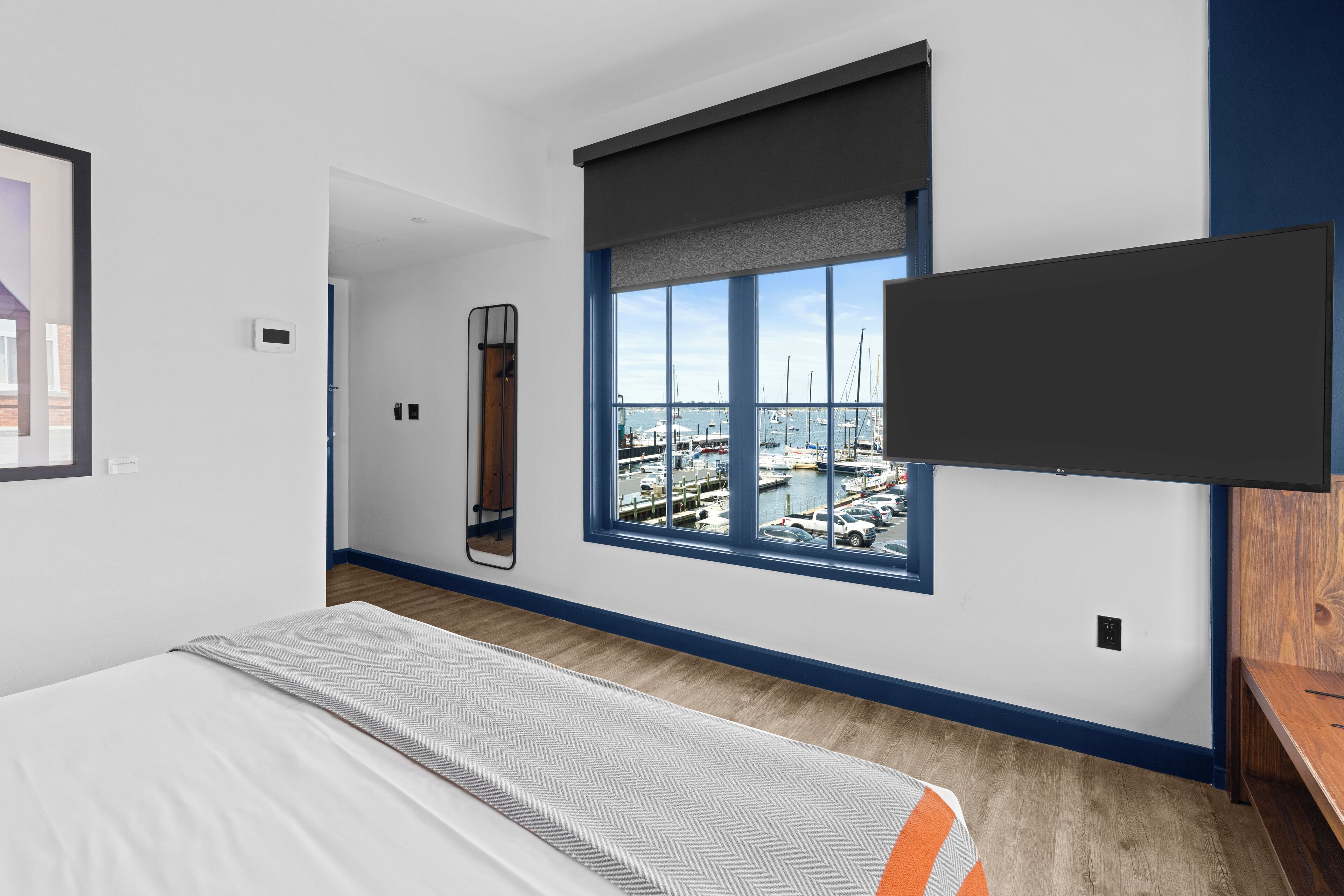 Une chambre d’hôtel moderne dotée d’une grande fenêtre donnant sur le port de plaisance, d’un téléviseur à écran plat fixé au mur et d’un lit recouvert d’une courtepointe à motifs.