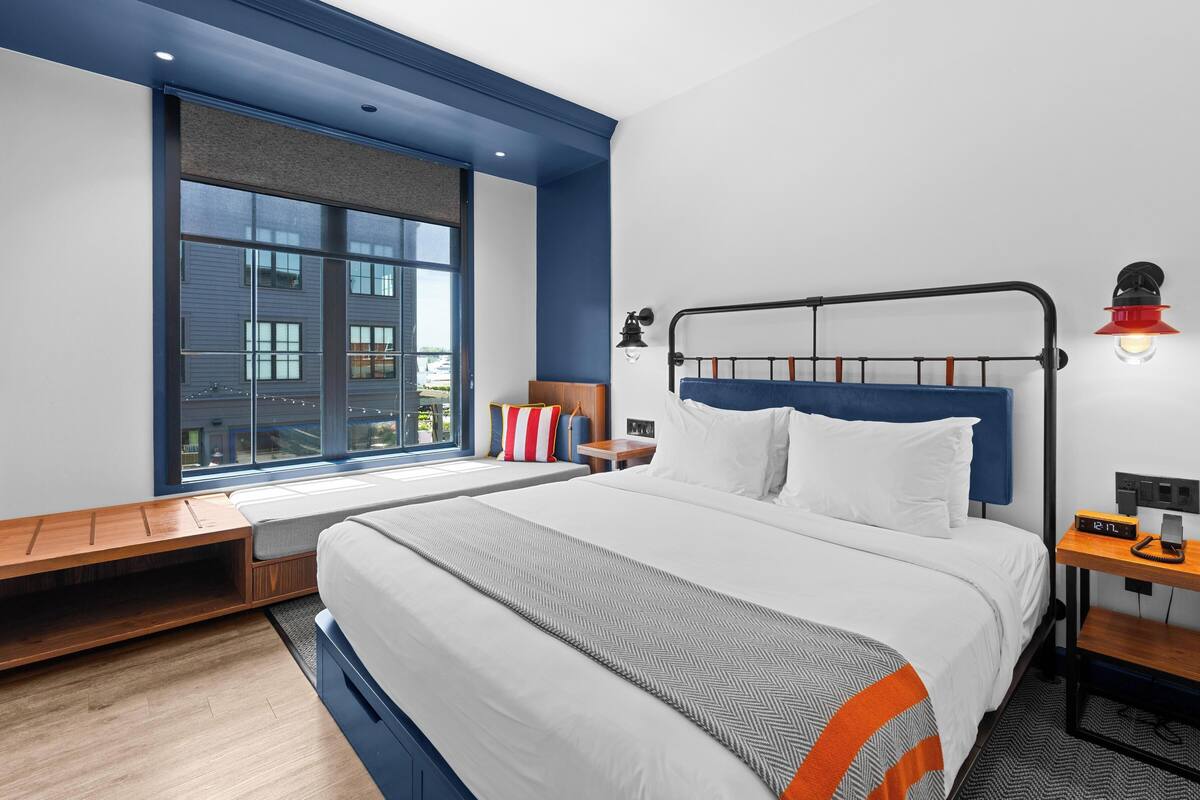 A modern hotel room with a large bed, a window with a view of buildings, a bedside table with a lamp, and a small wooden bench.
