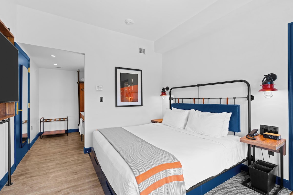 A hotel room with a bed, a bedside table with a lamp, a framed picture on the wall, and a wooden bench.