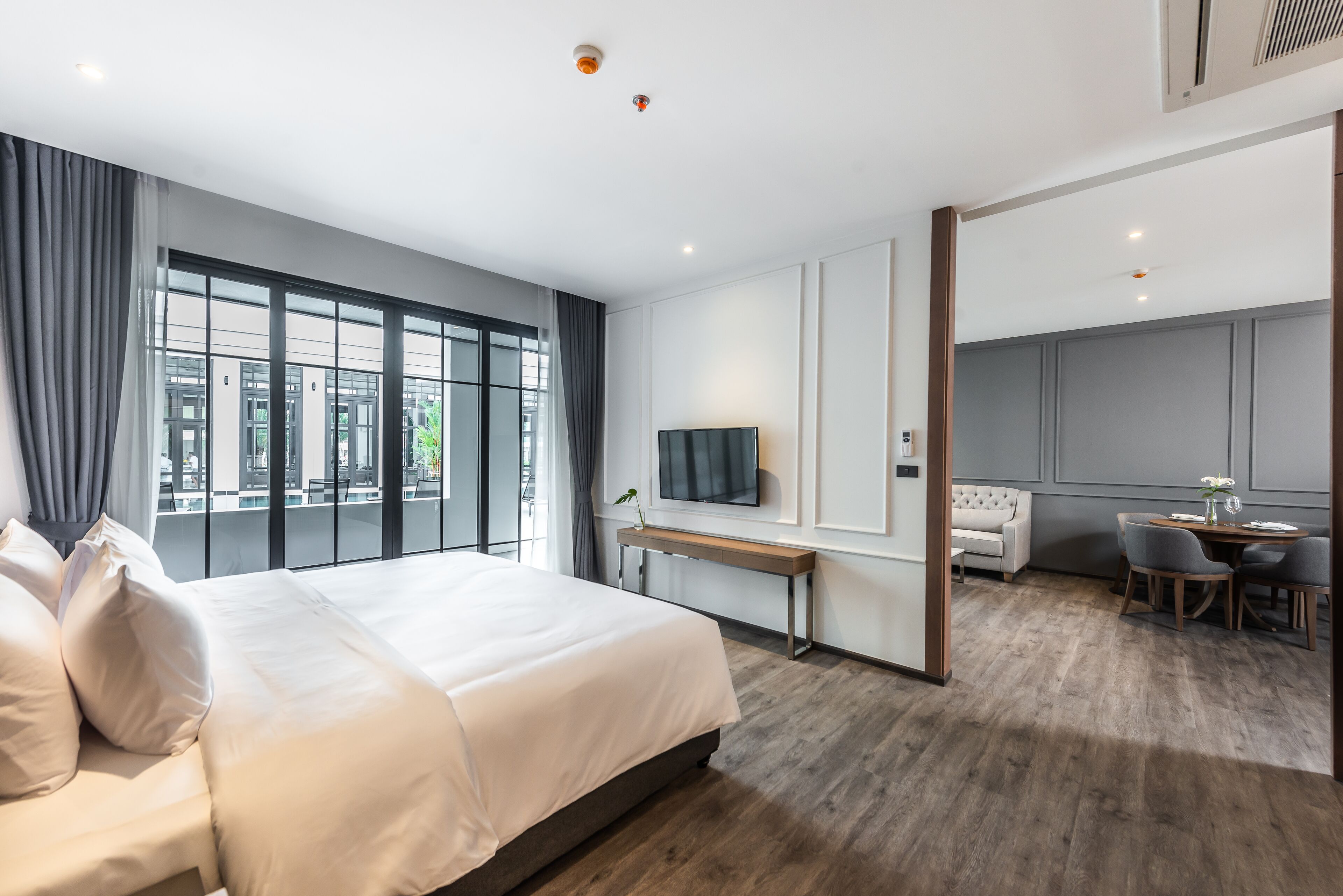 A modern hotel room with a large bed, a desk with a TV, a dining area, and large windows offering a city view.