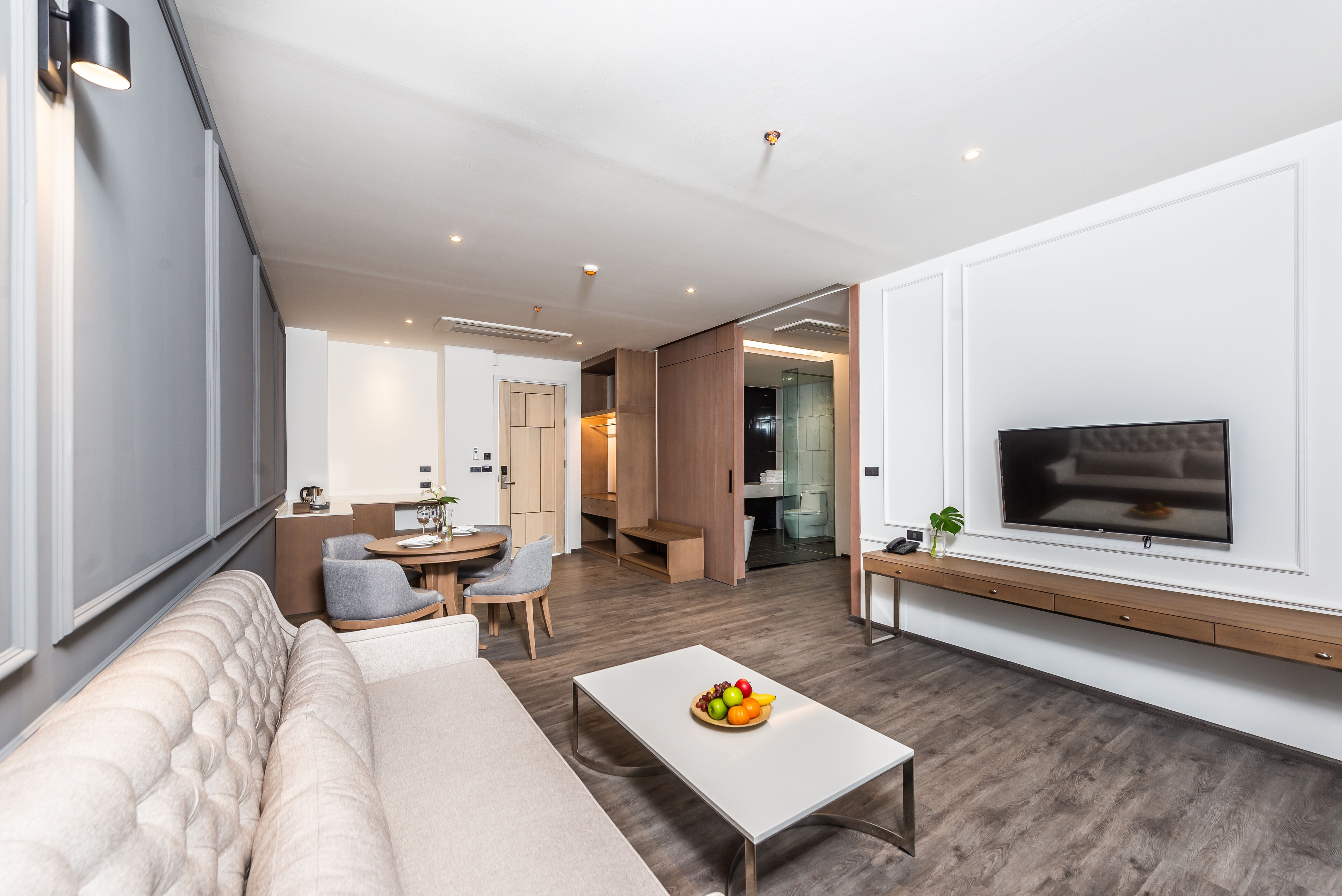 A modern living room with a sofa, coffee table, and TV. There is a dining area with chairs and a table, and a kitchen in the background.