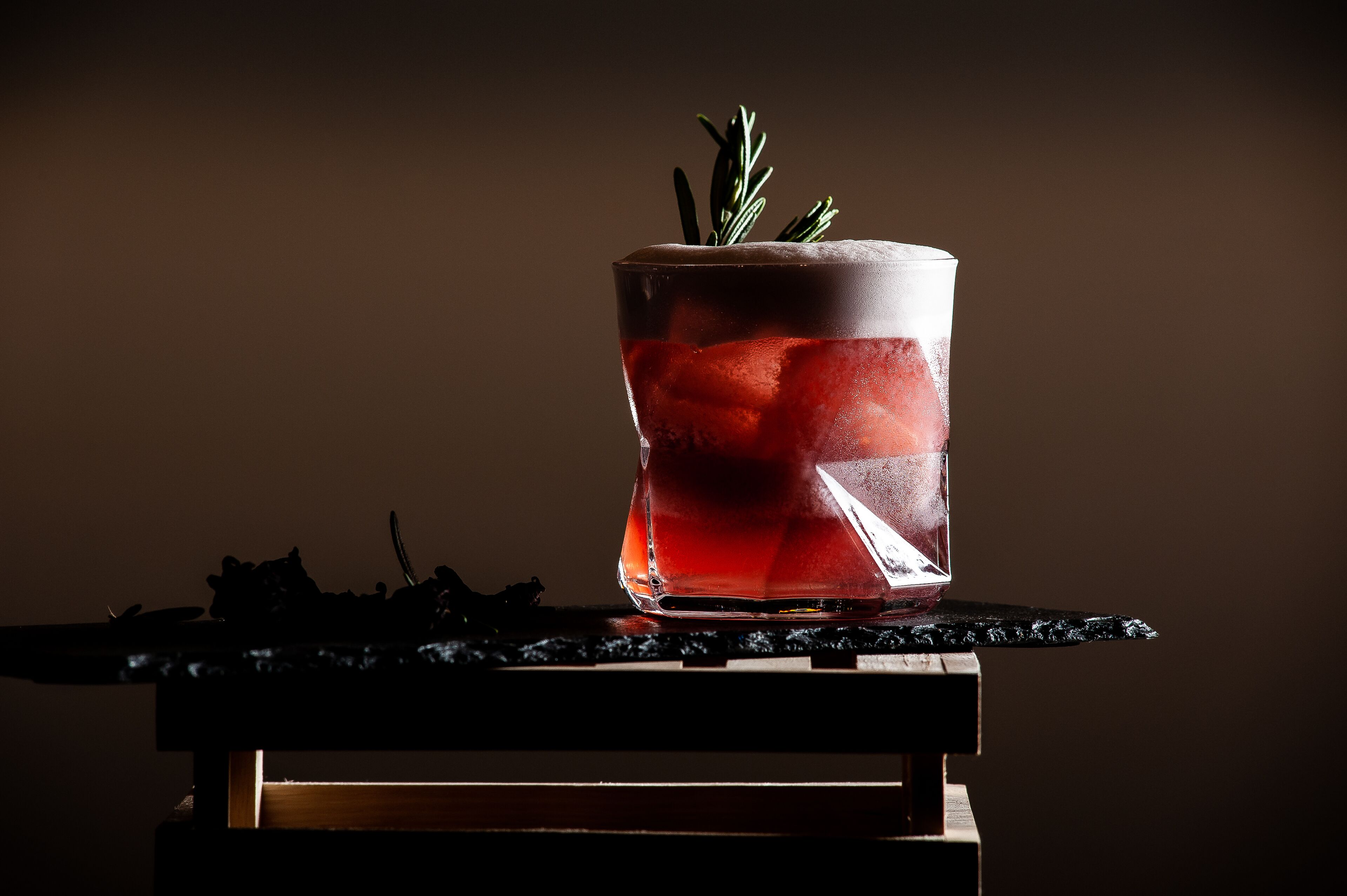 Ein Glas mit rotem Getränk und einem Rosmarinzweig auf einem dunklen Untersetzer.