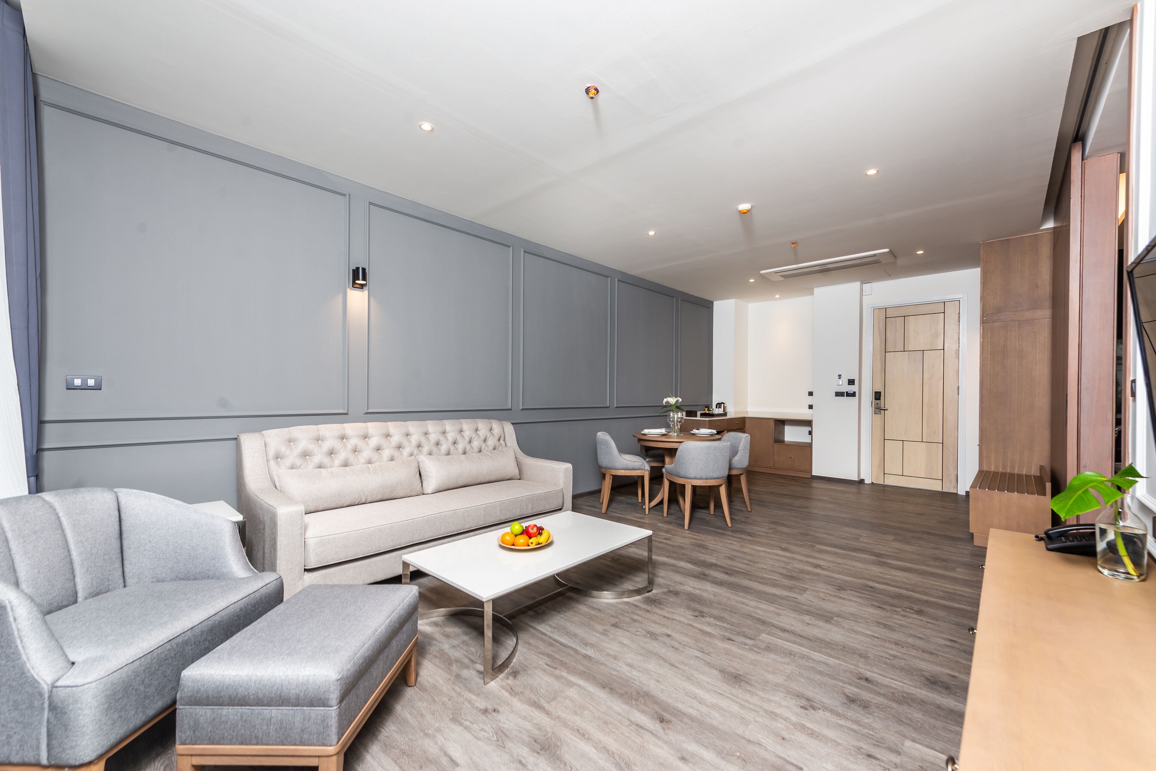 A modern living room with a grey sofa, a matching ottoman, and a coffee table with fruit. There is a dining area with a small table and chairs. The room has wooden flooring and light-colored walls.