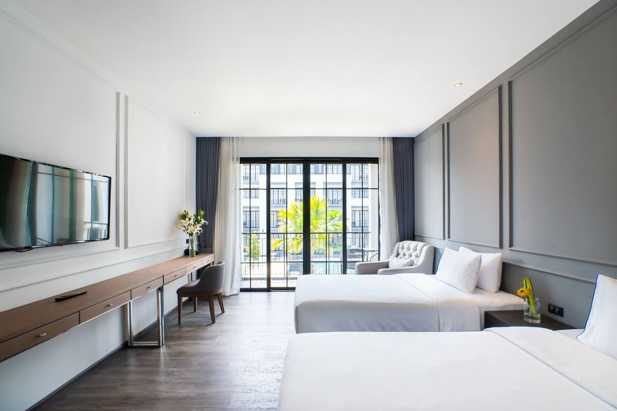A hotel room with a large bed, a desk, a chair, a TV, and a window with a view of buildings and trees.