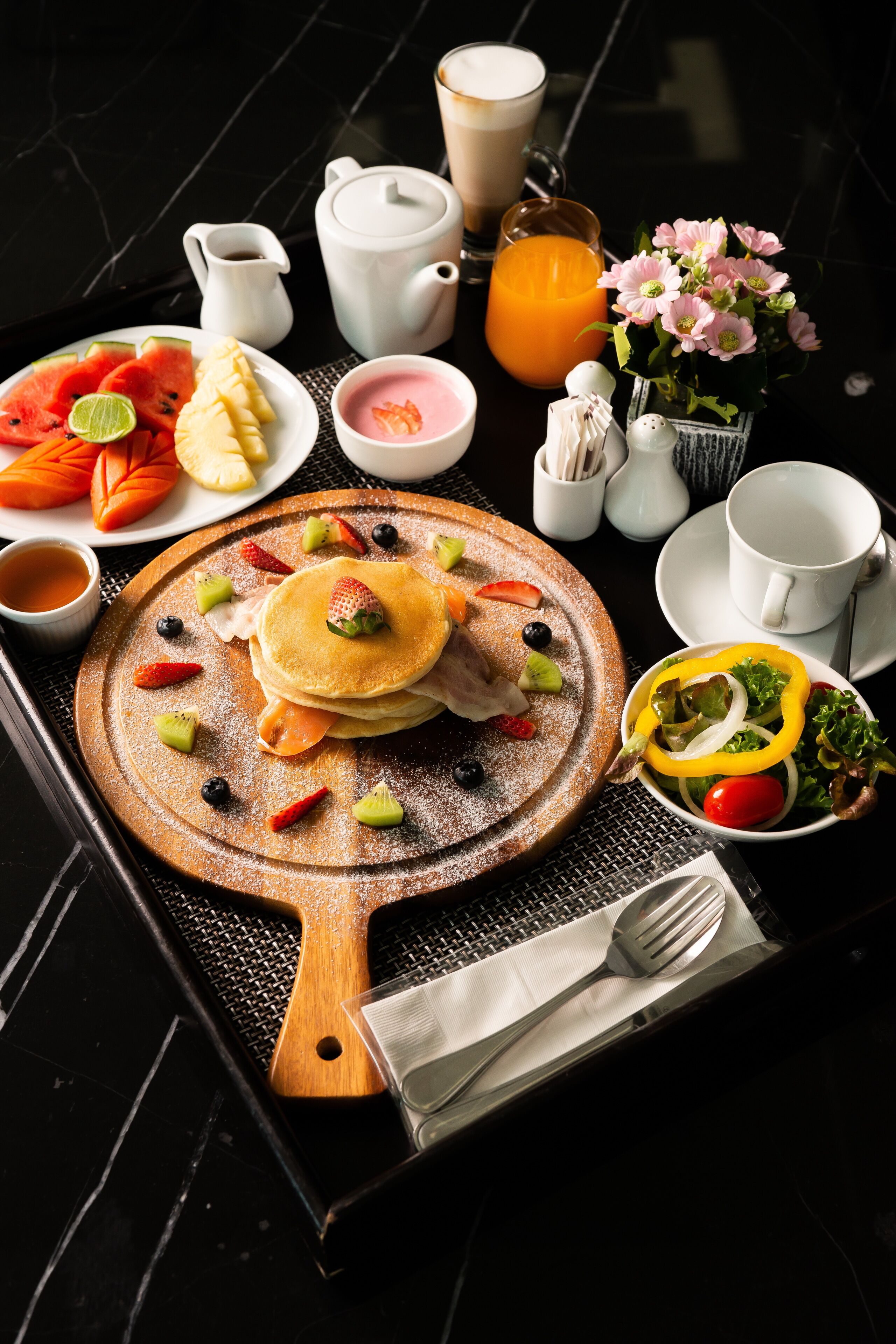 Desayuno con tortitas, frutas y bebidas sobre una mesa negra.