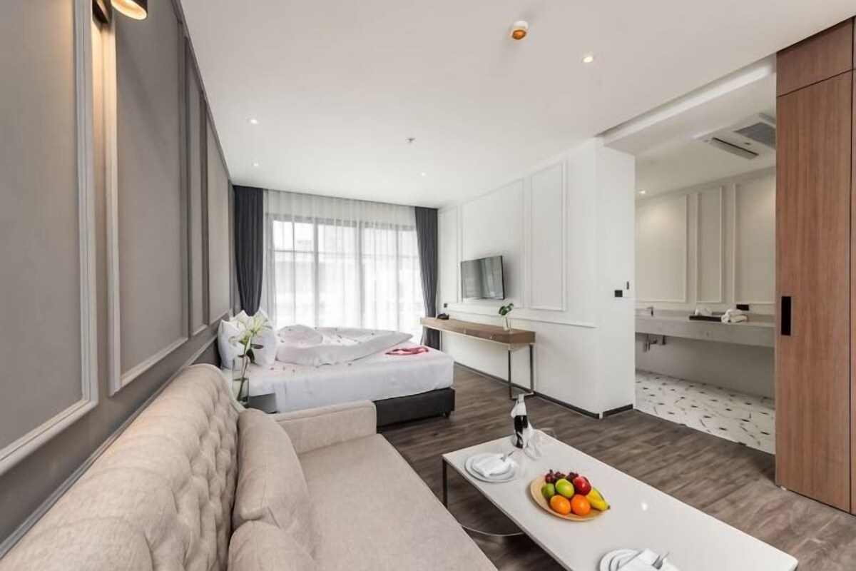 A modern hotel room with a large bed, a sitting area, a dining table with fruit, and a TV.