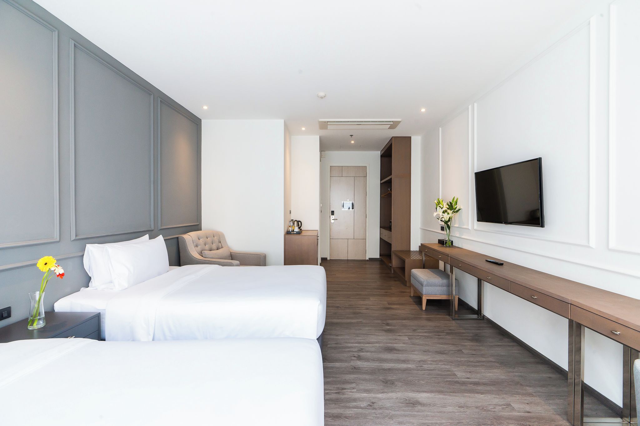 A hotel room with two beds, a desk, a chair, a TV, and a kitchenette in the background.