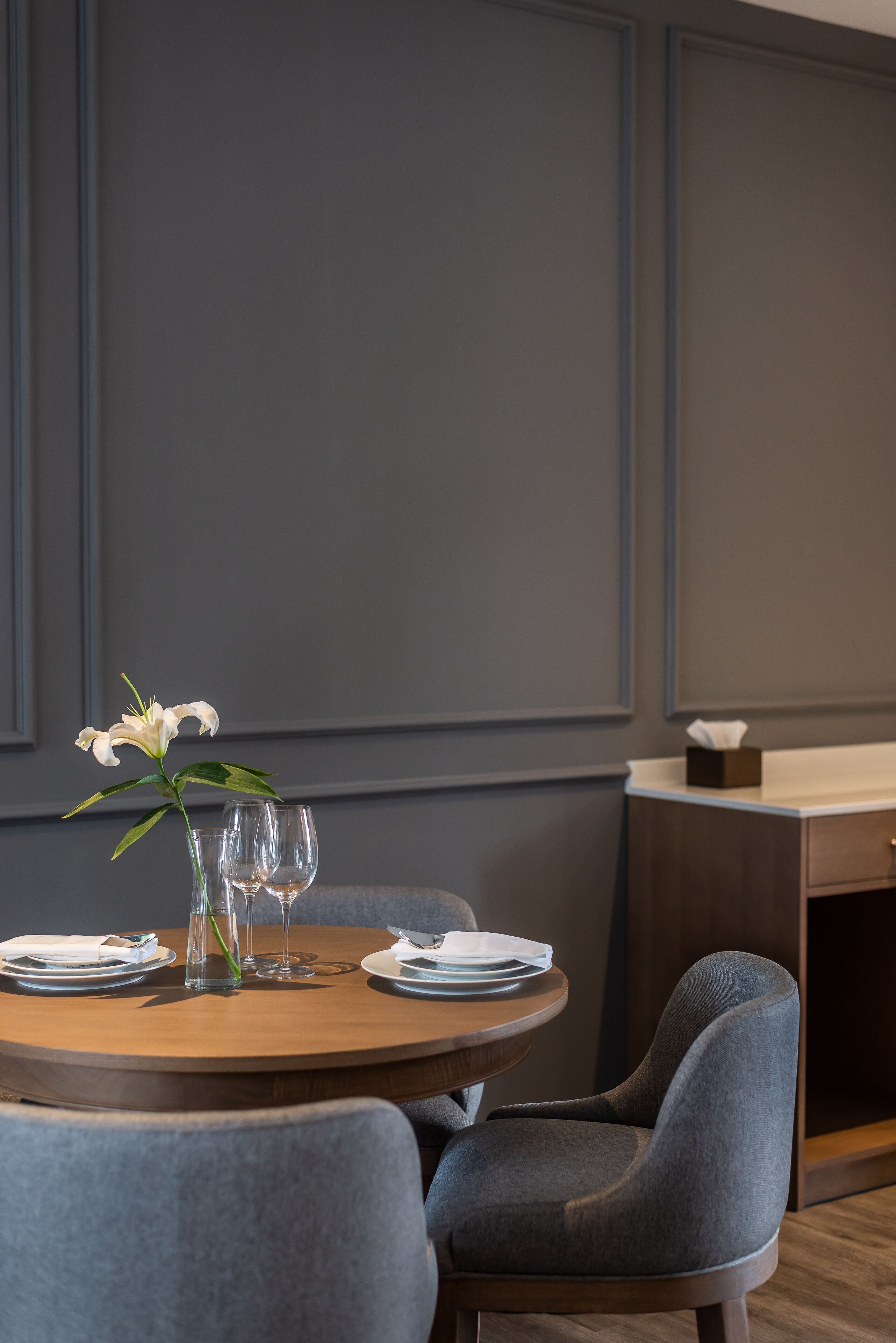A dining area with a round table set for two, a vase with a white flower, and a tissue box on a wooden counter.
