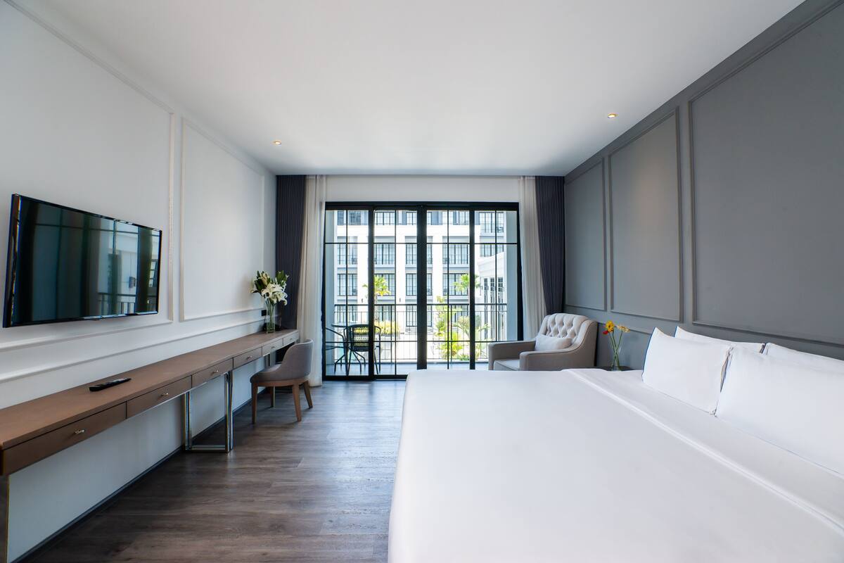 A modern hotel room with a large bed, a desk with a flat-screen TV, a chair, and a view of the city through large windows.