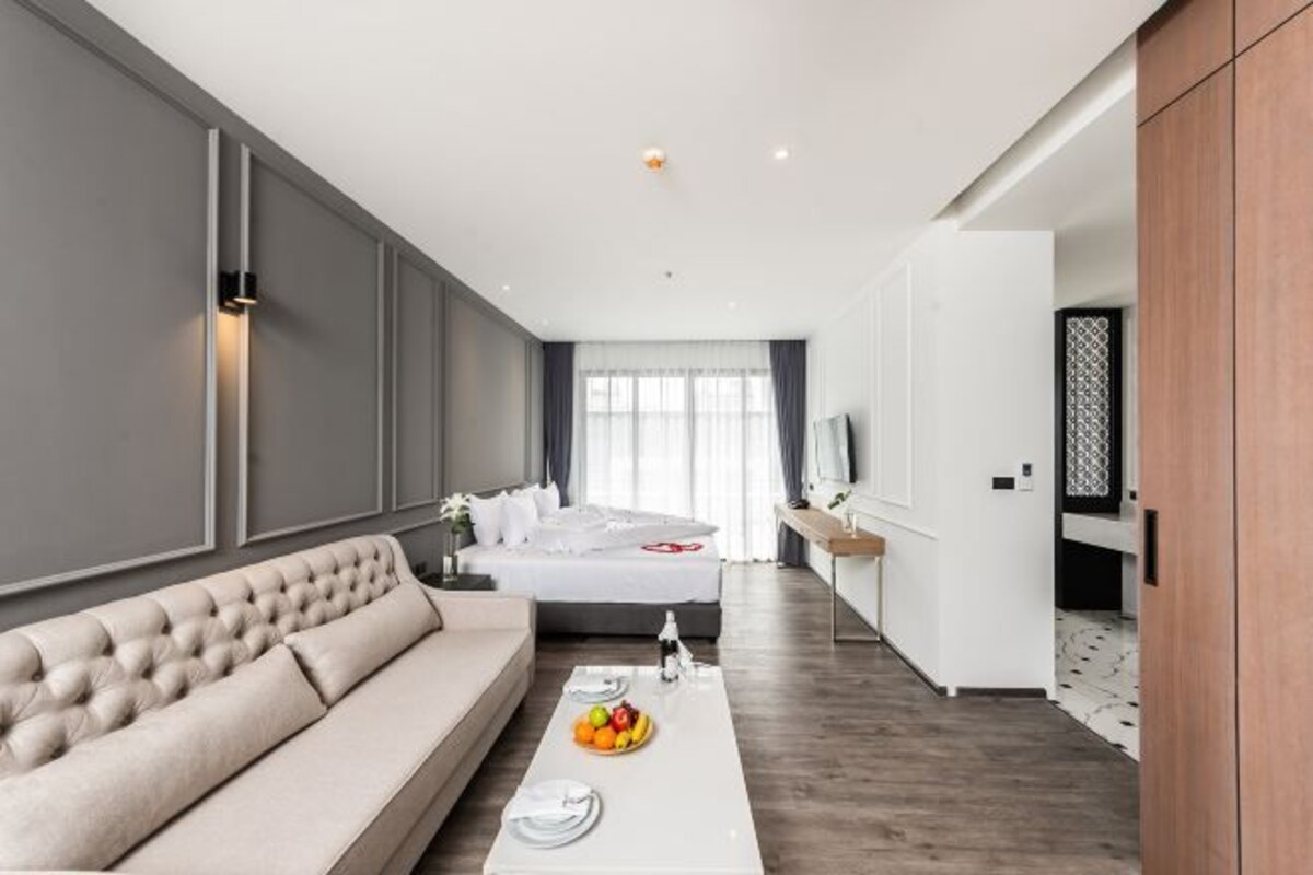 A modern hotel room with a large bed, a sofa, a dining table with fruit, and a TV.