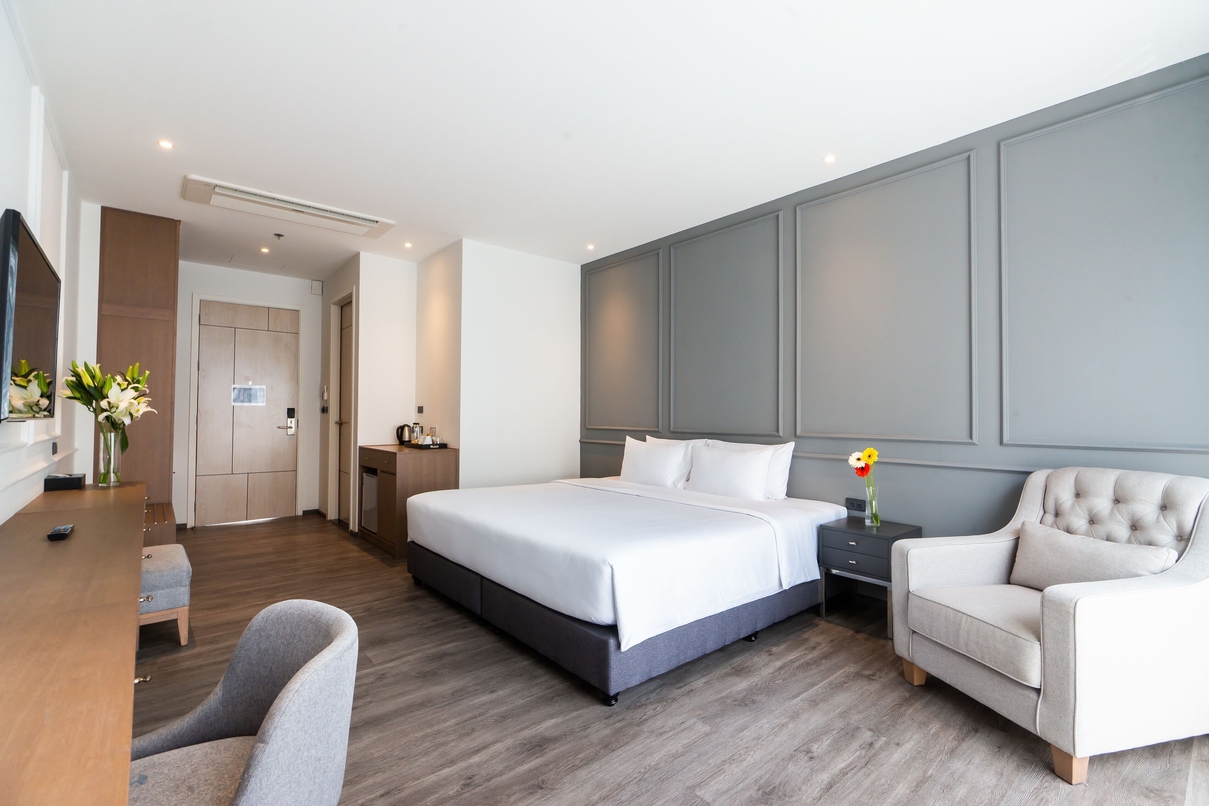 A modern hotel room with a large bed, a bedside table, a chair, and a desk with a vase of flowers.