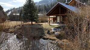 A cabin with a patio, a small stream, and a forested backdrop.
