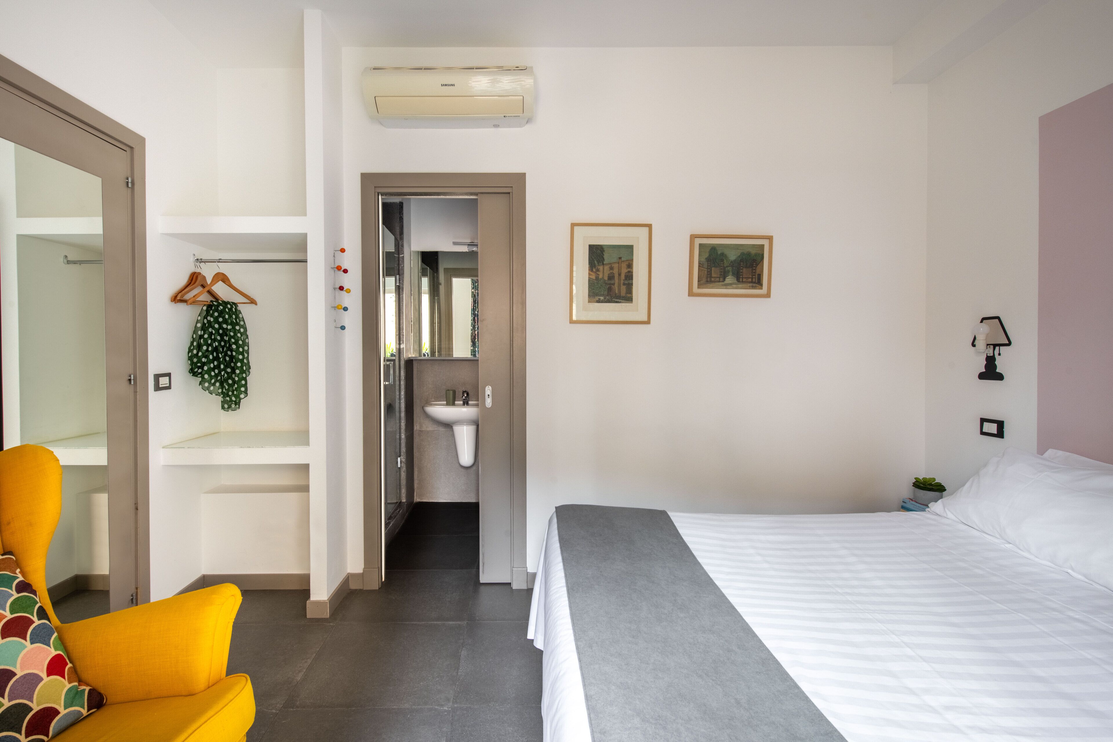 A hotel room with a bed, a chair, a wardrobe, a sink, and two framed pictures on the wall.