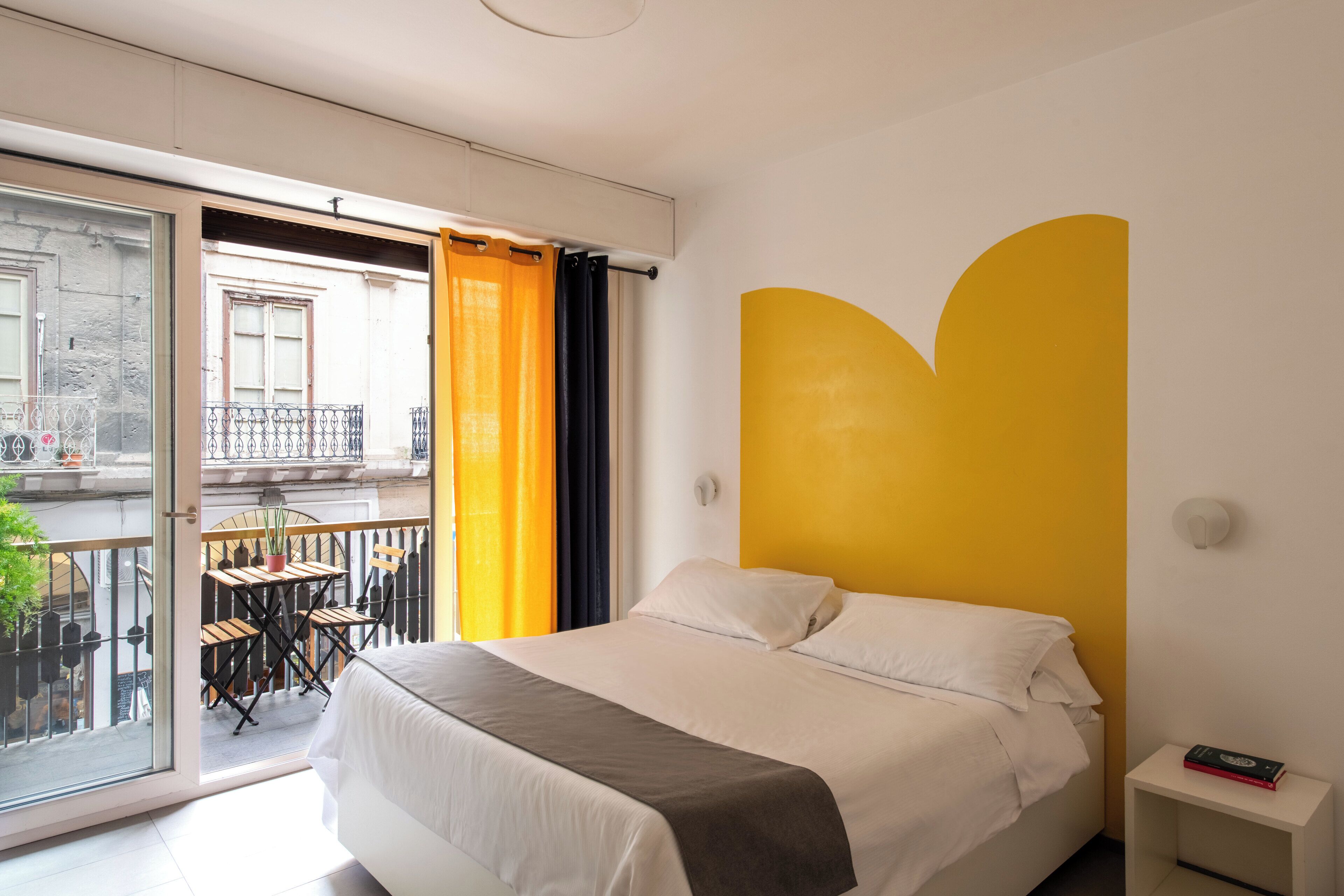 A bedroom with a bed, a nightstand, a window with curtains, a balcony with a table and chairs, and a yellow accent wall.