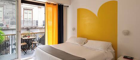 A bedroom with a bed, a nightstand, a window with curtains, a balcony with a table and chairs, and a yellow accent wall.