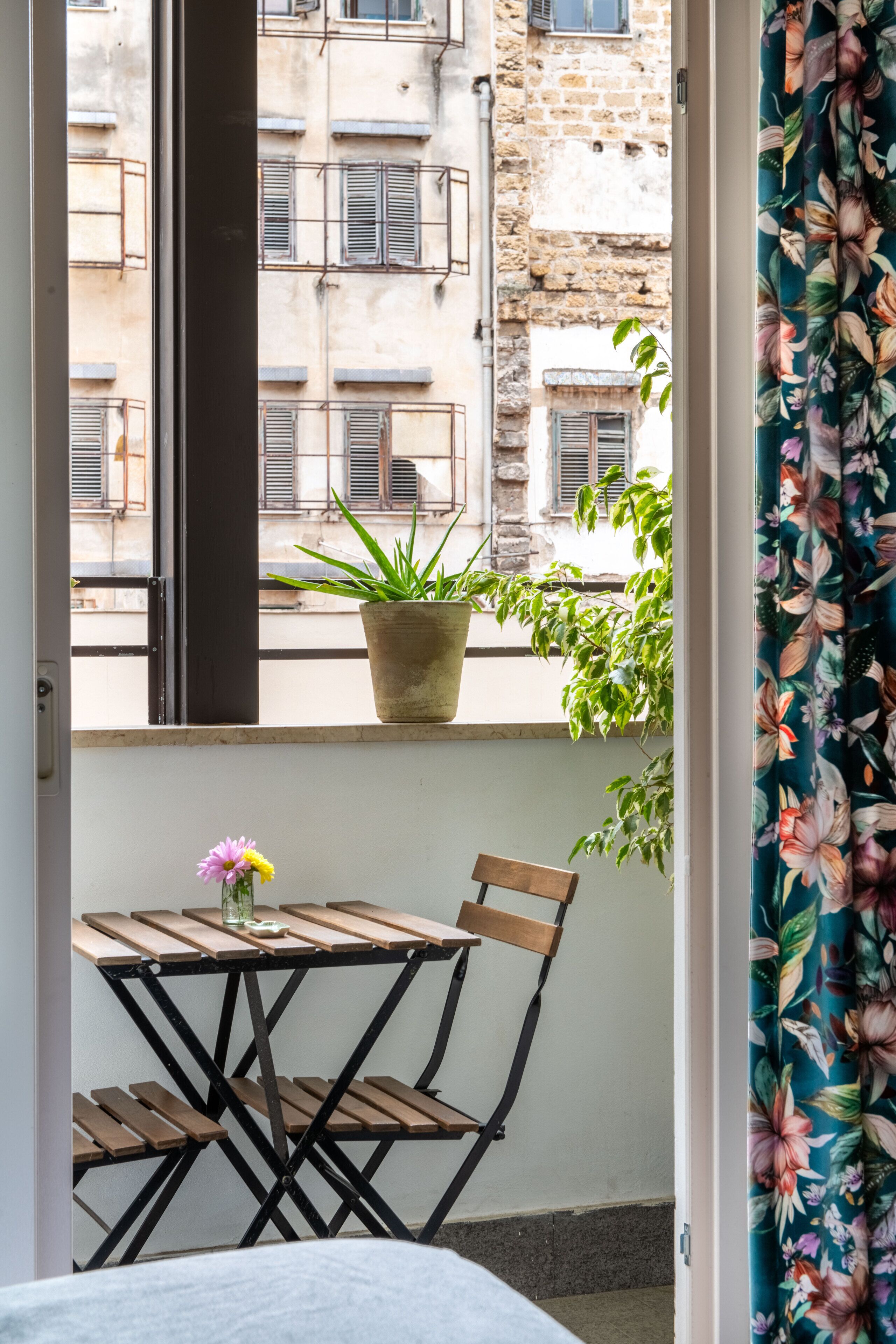A balcony with a wooden table and chairs, a potted plant, and a view of a building with stone walls and windows.