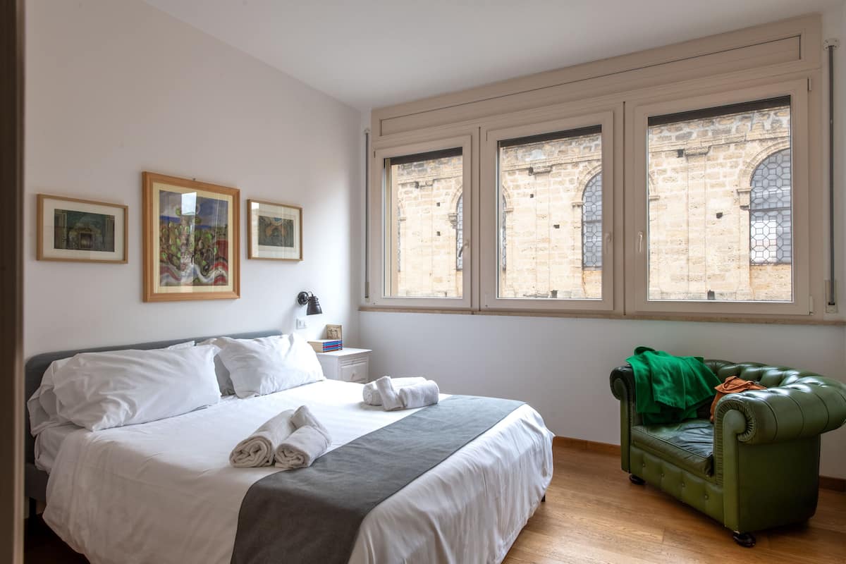 A hotel room with a bed, a green armchair, and three windows with a view of a stone building.