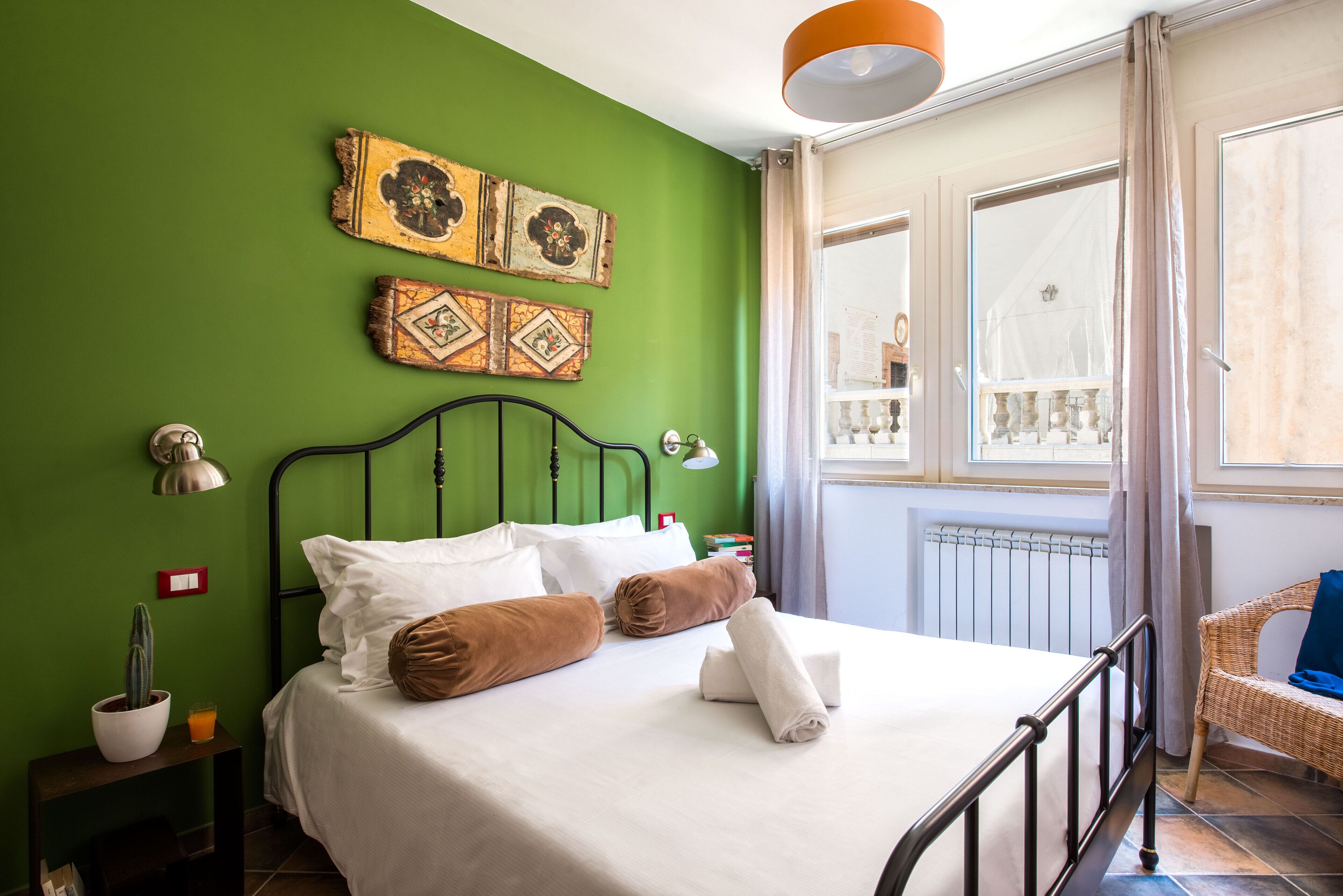 A neatly made bed with white linens and brown pillows, a green wall with decorative tiles, a wicker chair, and a window with curtains.