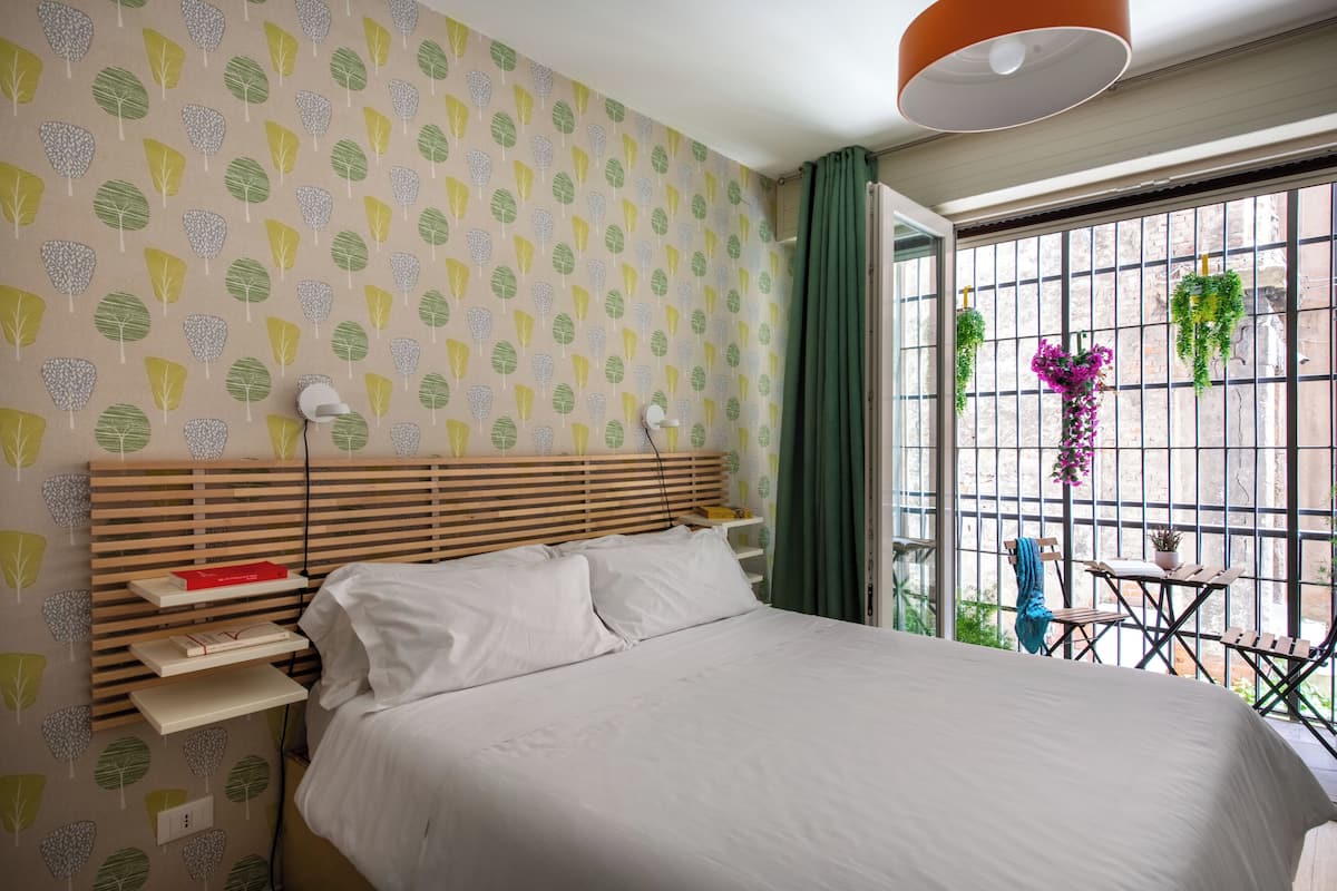 A bedroom with a bed, bedside tables, a window with a view of greenery, and a hanging plant.