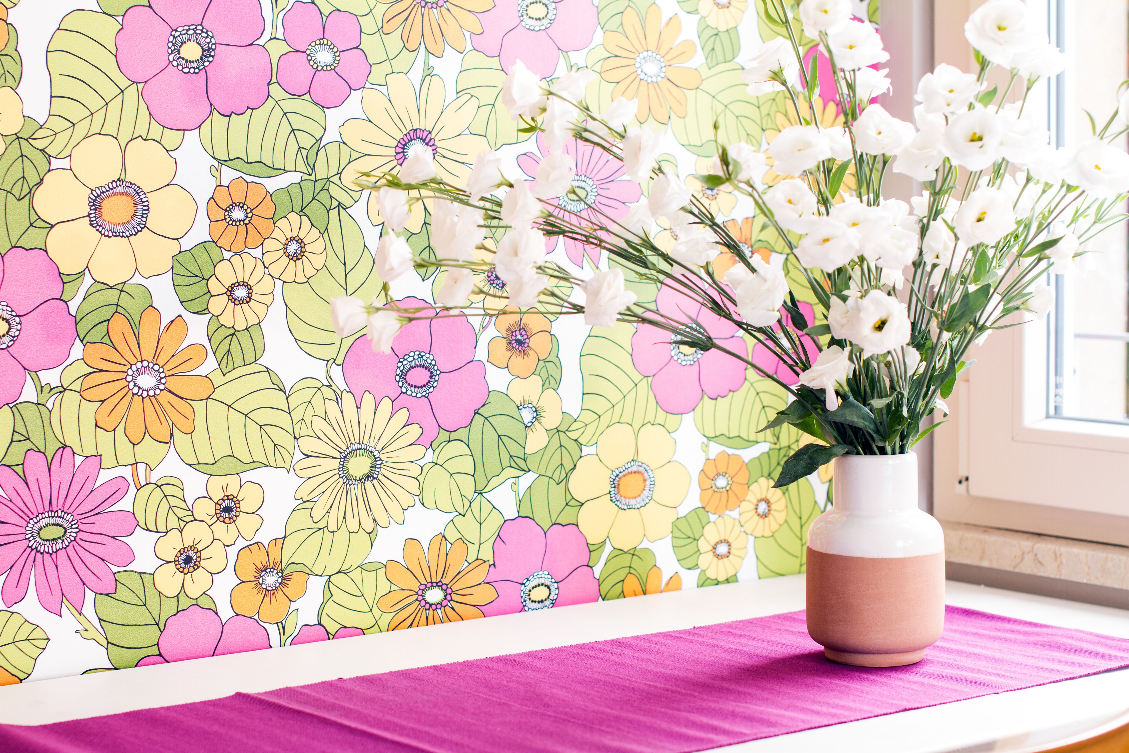 Una habitación con papel tapiz floral, un jarrón con flores blancas y un mantel rosa.