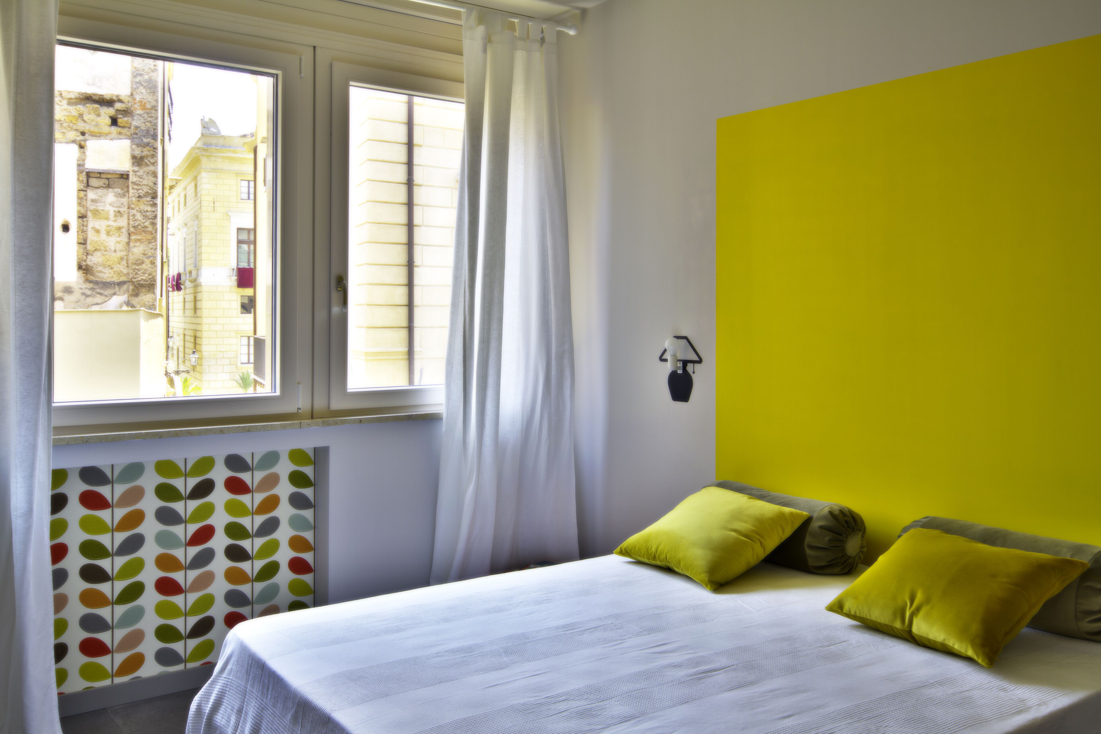 Un dormitorio con una cama, una ventana con cortinas, un radiador con motivos de hojas coloridas y una pared de color amarillo brillante.
