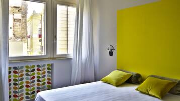 Un dormitorio con una cama, una ventana con cortinas, un radiador con motivos de hojas coloridas y una pared de color amarillo brillante.