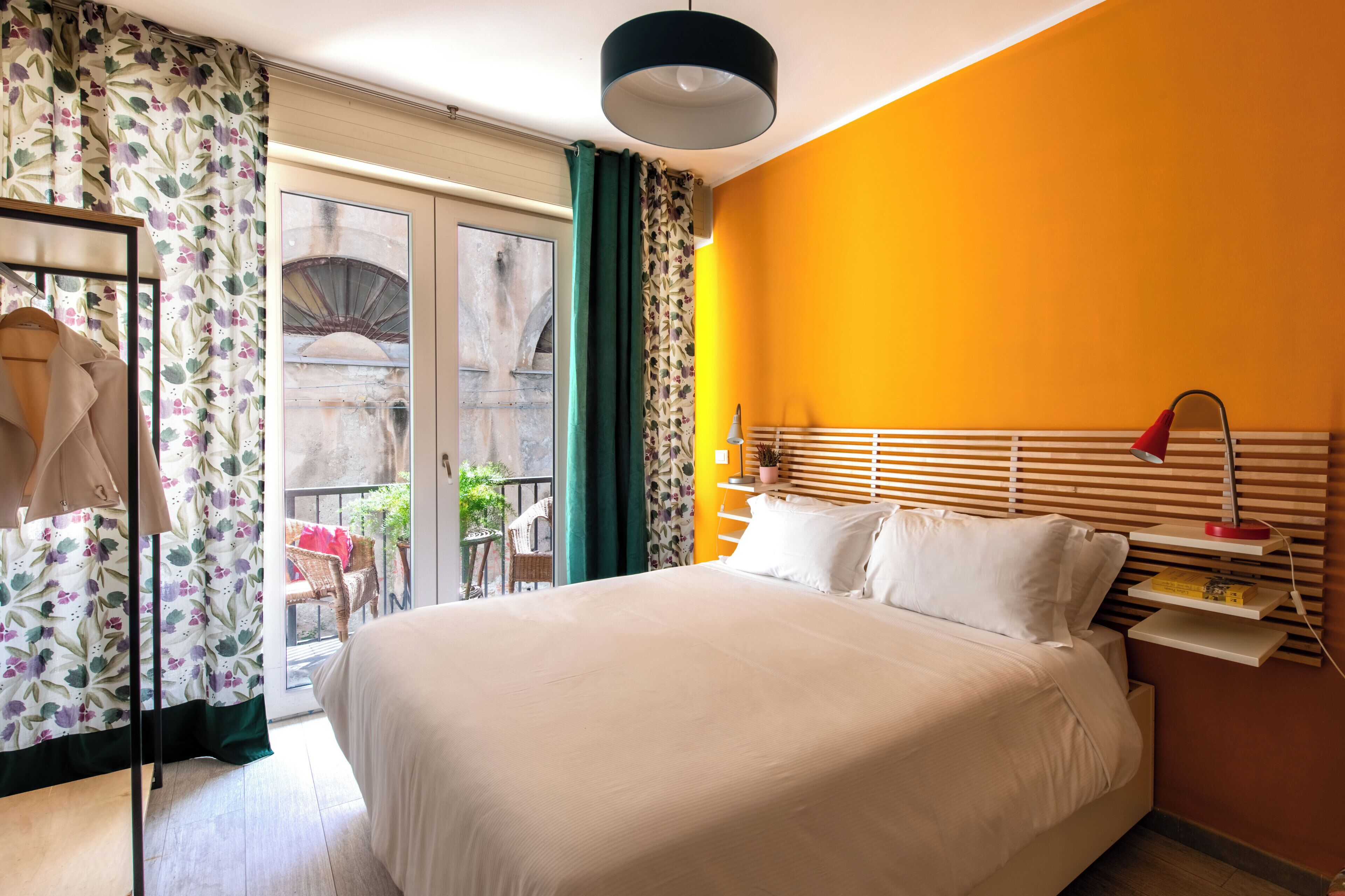 Quarto de hotel com uma cama grande, cabeceira de madeira, mesa de cabeceira com abajur e vista para uma varanda com cadeiras e plantas.