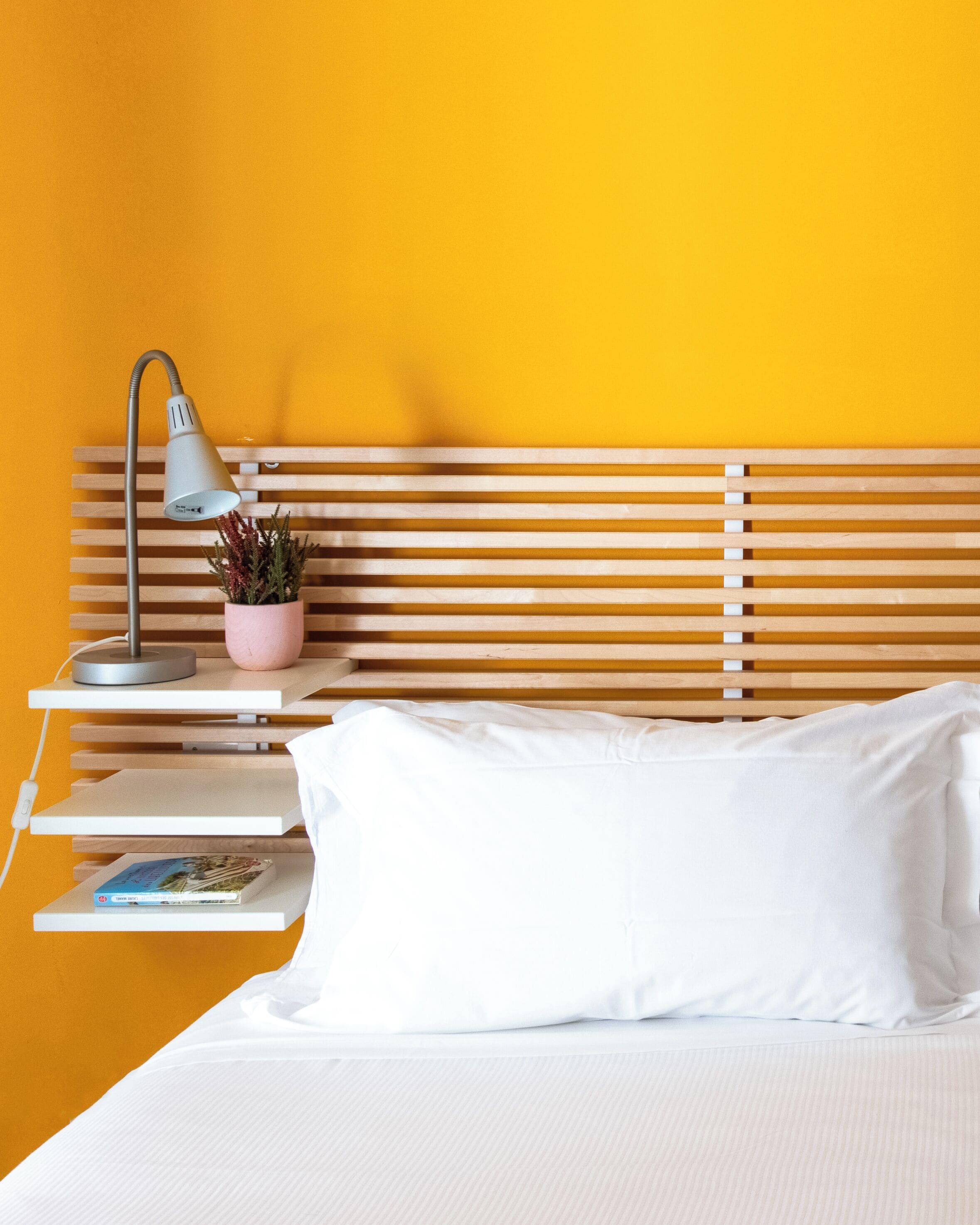 Una cama con ropa de cama blanca, una lámpara de noche, una planta rosa y un libro en una estantería.