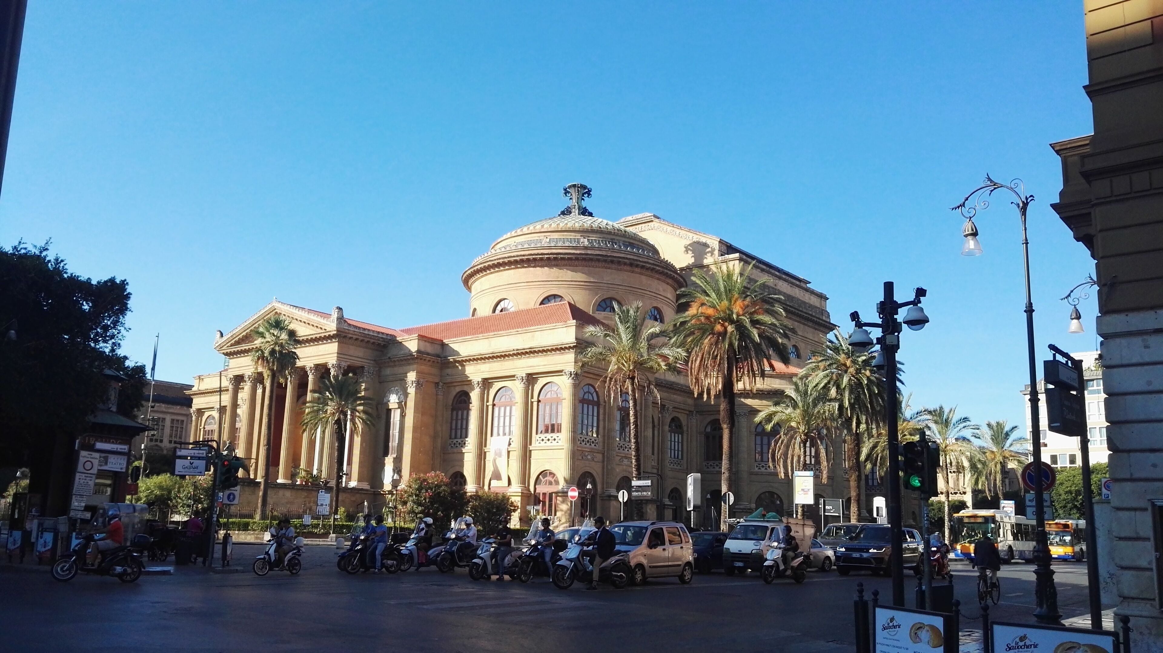 A historic building with a dome and columns, surrounded by palm trees and parked vehicles.