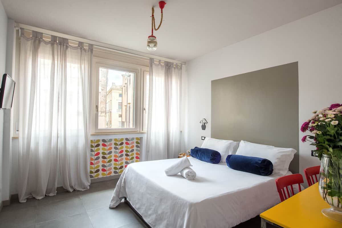 A hotel room with a bed, a window with sheer curtains, a yellow table, and a vase with flowers.